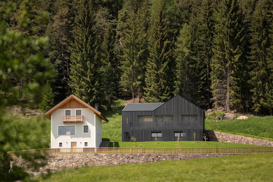 Two modern houses, one white with a wooden roof and balcony, and one dark grey with a sleek design, sit side by side on green grass in front of a dense forest of tall pine trees.