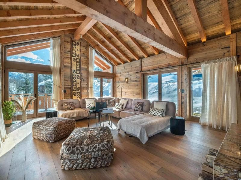 Spacious mountain cabin living room with large windows, wood beams, a sectional sofa with patterned pillows and ottomans, plants, and views of snowy peaks outside. Natural light fills the cozy, rustic space.