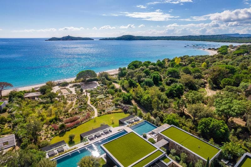 Aerial view of a coastal resort with modern buildings, green rooftops, gardens, and two swimming pools near a sandy beach and clear blue sea, bordered by lush trees and distant hills under a partly cloudy sky.