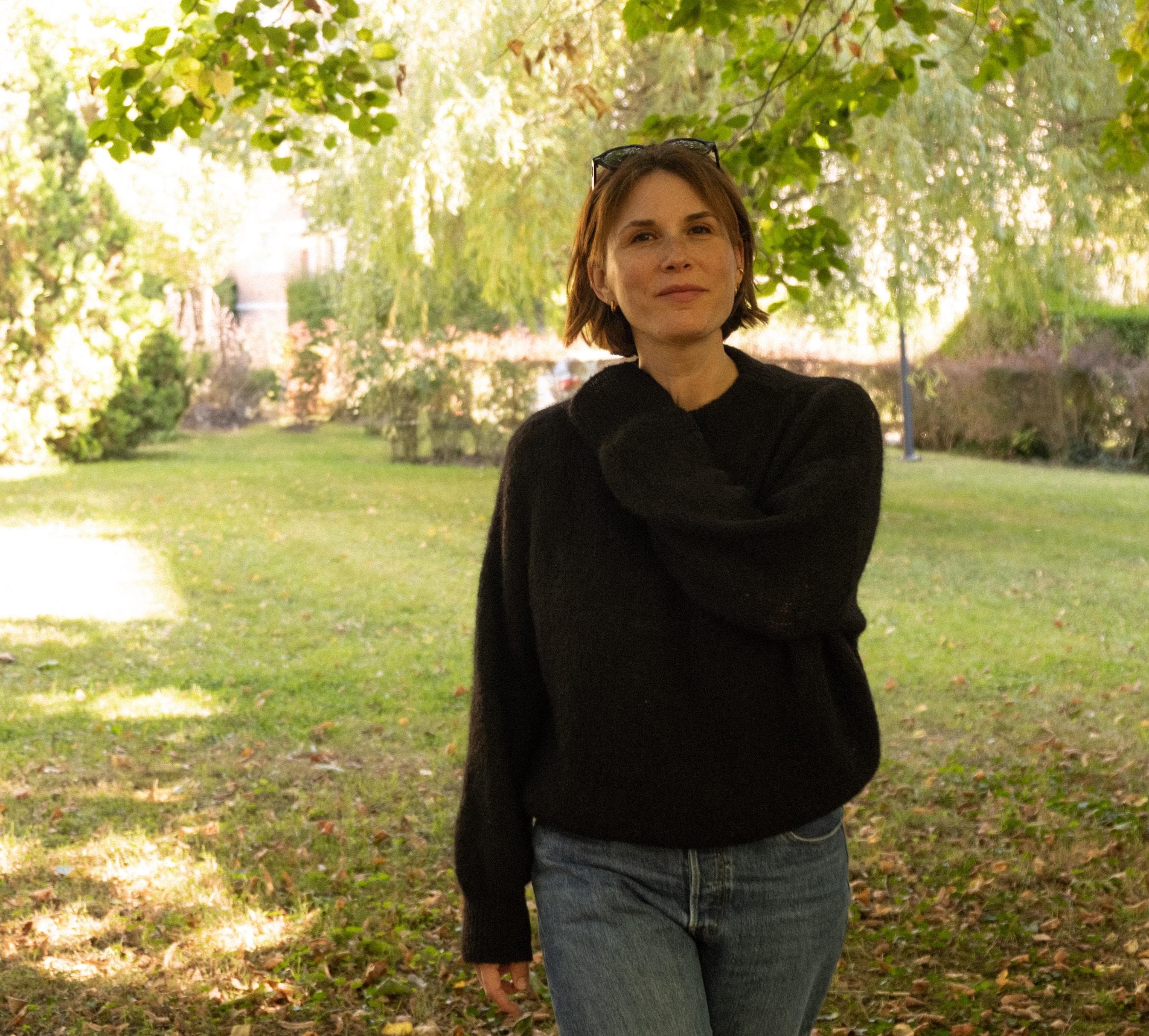 A person with short brown hair, wearing a dark sweater and blue jeans, stands outdoors in a grassy, sunlit park surrounded by trees and greenery.