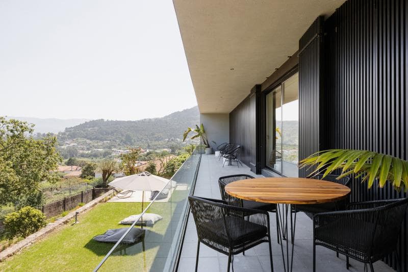 Un balcon moderne avec une table ronde en bois et des chaises noires donne sur une cour herbeuse avec des chaises longues et un parasol, avec des montagnes et des arbres visibles au loin.