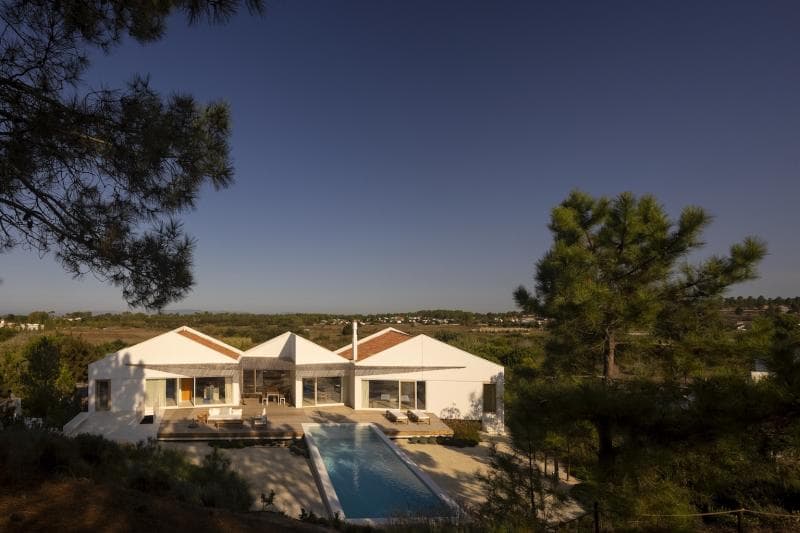A modern white villa with large windows and a central swimming pool, surrounded by trees and greenery, under a clear blue sky at sunset.