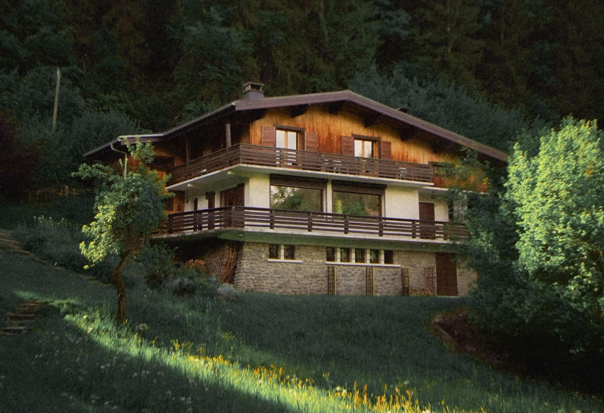 A rustic wooden chalet with stone foundation sits on a grassy hillside, surrounded by trees and lush greenery, with sunlight filtering through onto the lawn.