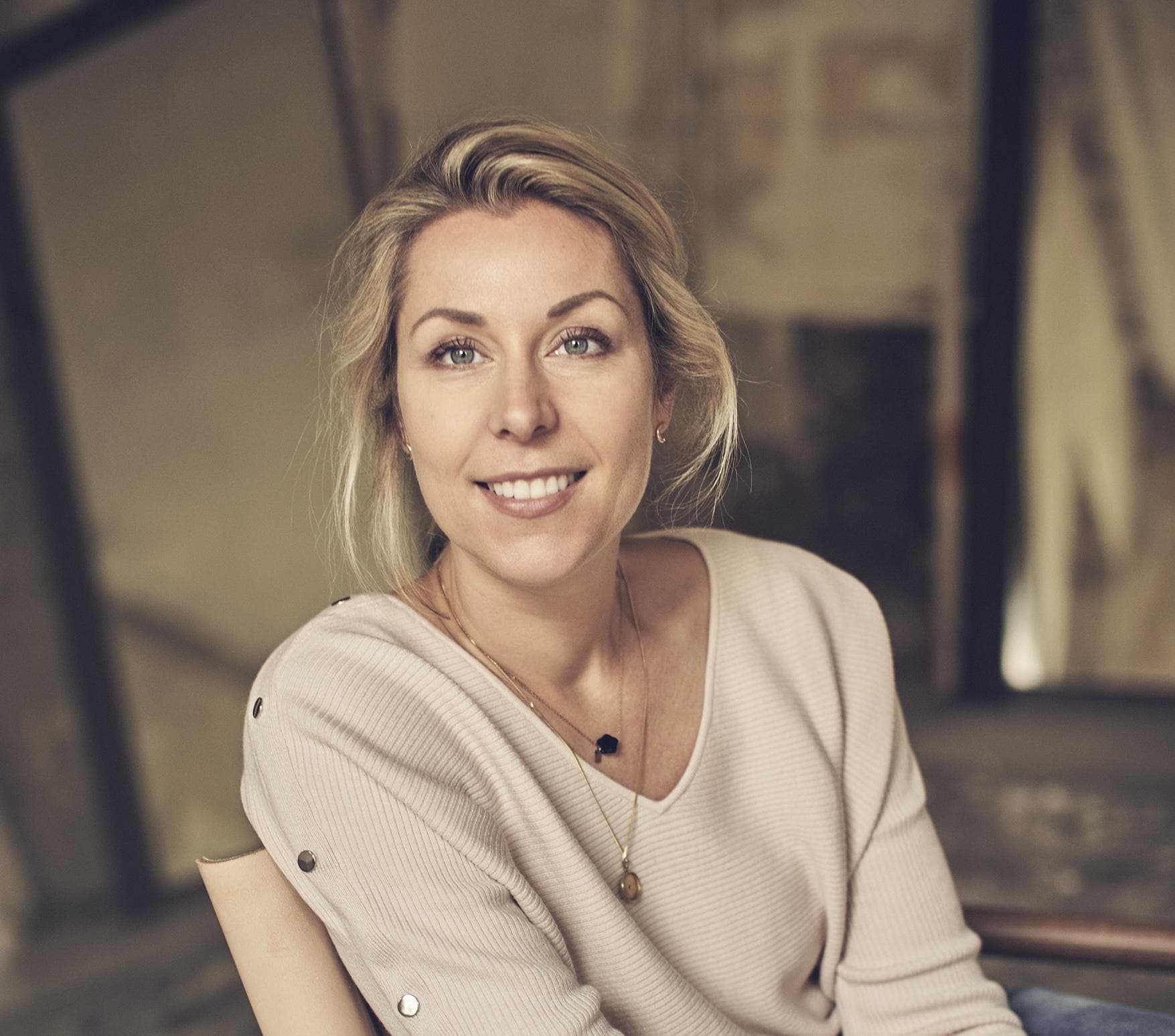 A woman with blonde hair tied back, wearing a light beige sweater with button details on the sleeve and a gold necklace, sits smiling in a softly lit indoor setting with a blurred, rustic background.
