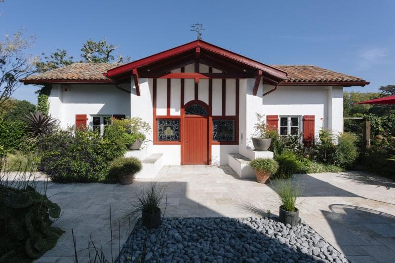 A small white house with red trim, red shutters, and a red door. The house has a tile roof and is surrounded by green plants and potted shrubs, with a stone patio and blue sky in the background.