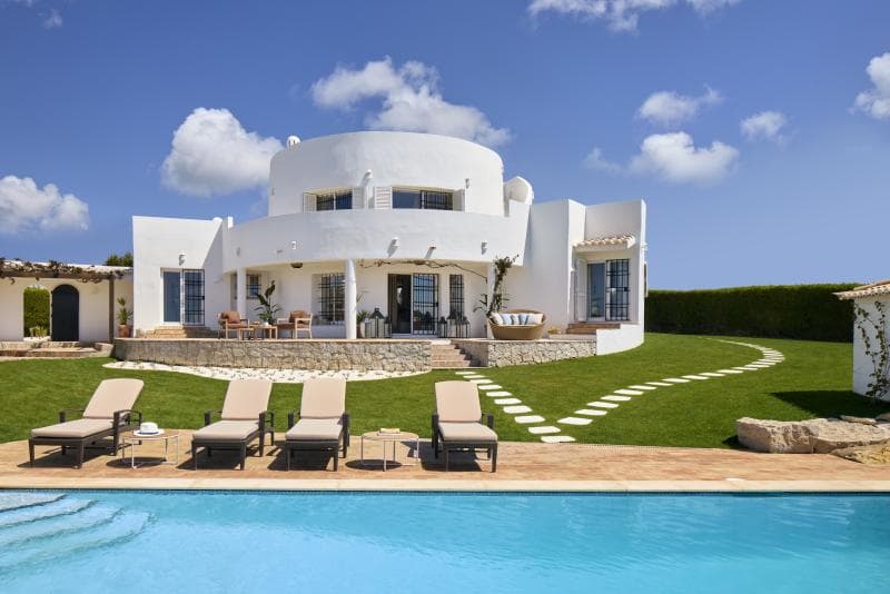 A modern white villa with rounded architecture, large windows, and a spacious patio overlooks a swimming pool with four lounge chairs on a sunny day. The surrounding lawn is neatly landscaped with stepping stones.
