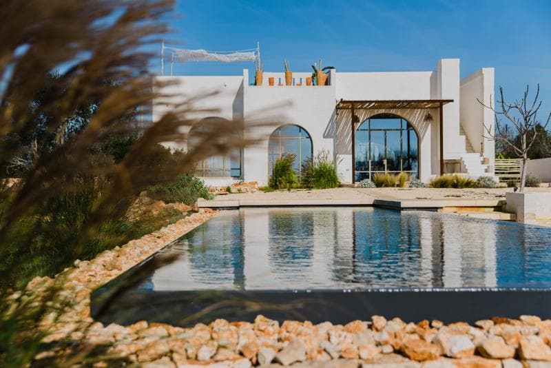 Modern white house with large windows and a shaded patio, surrounded by a garden and rocks, reflected in a rectangular pool under a clear blue sky. Some grasses are blurred in the foreground.