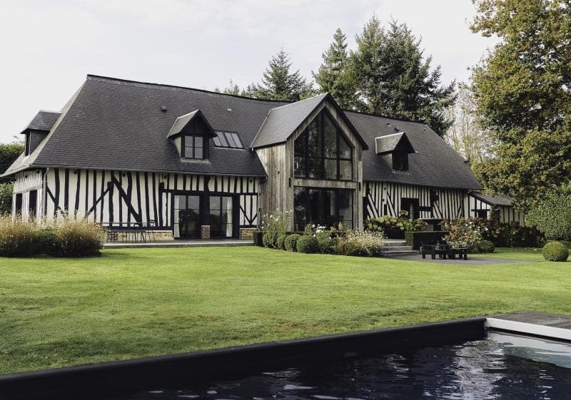A large, modern Tudor-style house with black timber framing and white walls sits behind a neatly manicured lawn, garden, and a swimming pool, surrounded by tall trees.