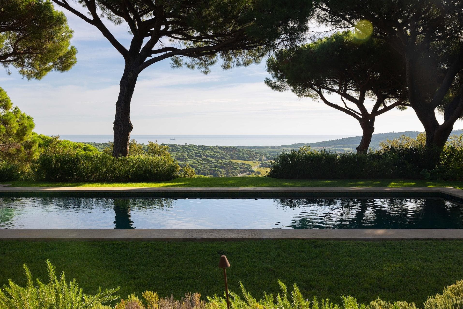 A serene swimming pool reflects tall trees, with lush greenery and rolling hills in the background, and a distant view of the sea under a partly cloudy sky.
