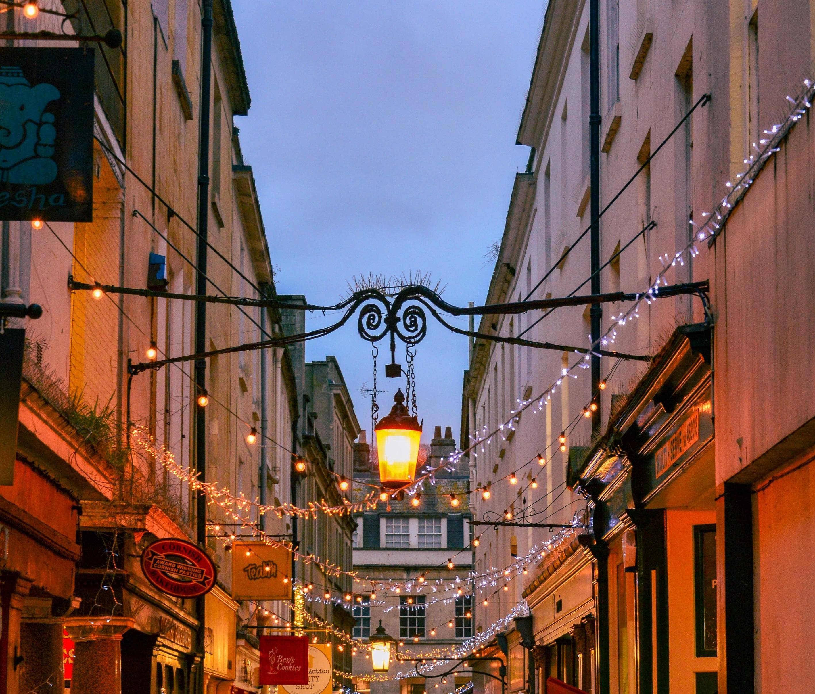 A narrow street lined with shops is decorated with string lights and a central glowing lantern, creating a festive, cozy atmosphere at dusk. The sky is cloudy, and buildings frame the lively scene.
