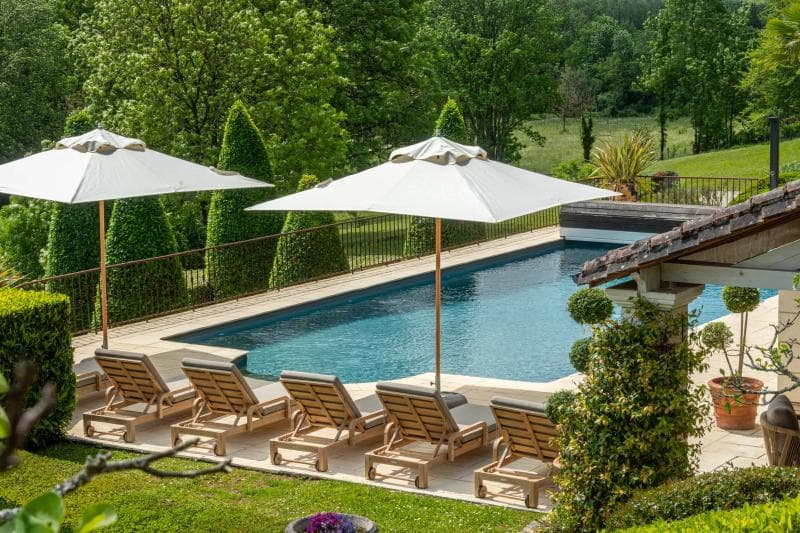 A rectangular outdoor swimming pool with six wooden lounge chairs and two large white umbrellas, surrounded by greenery and trees in a landscaped garden.