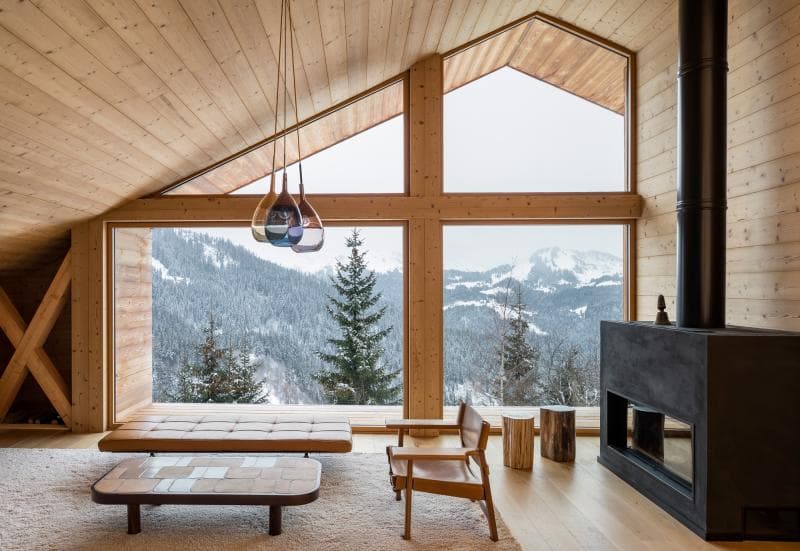 A modern wooden cabin interior with large windows overlooking a snowy mountain landscape, featuring a cozy seating area, minimalist furniture, a black fireplace, and natural wood finishes.