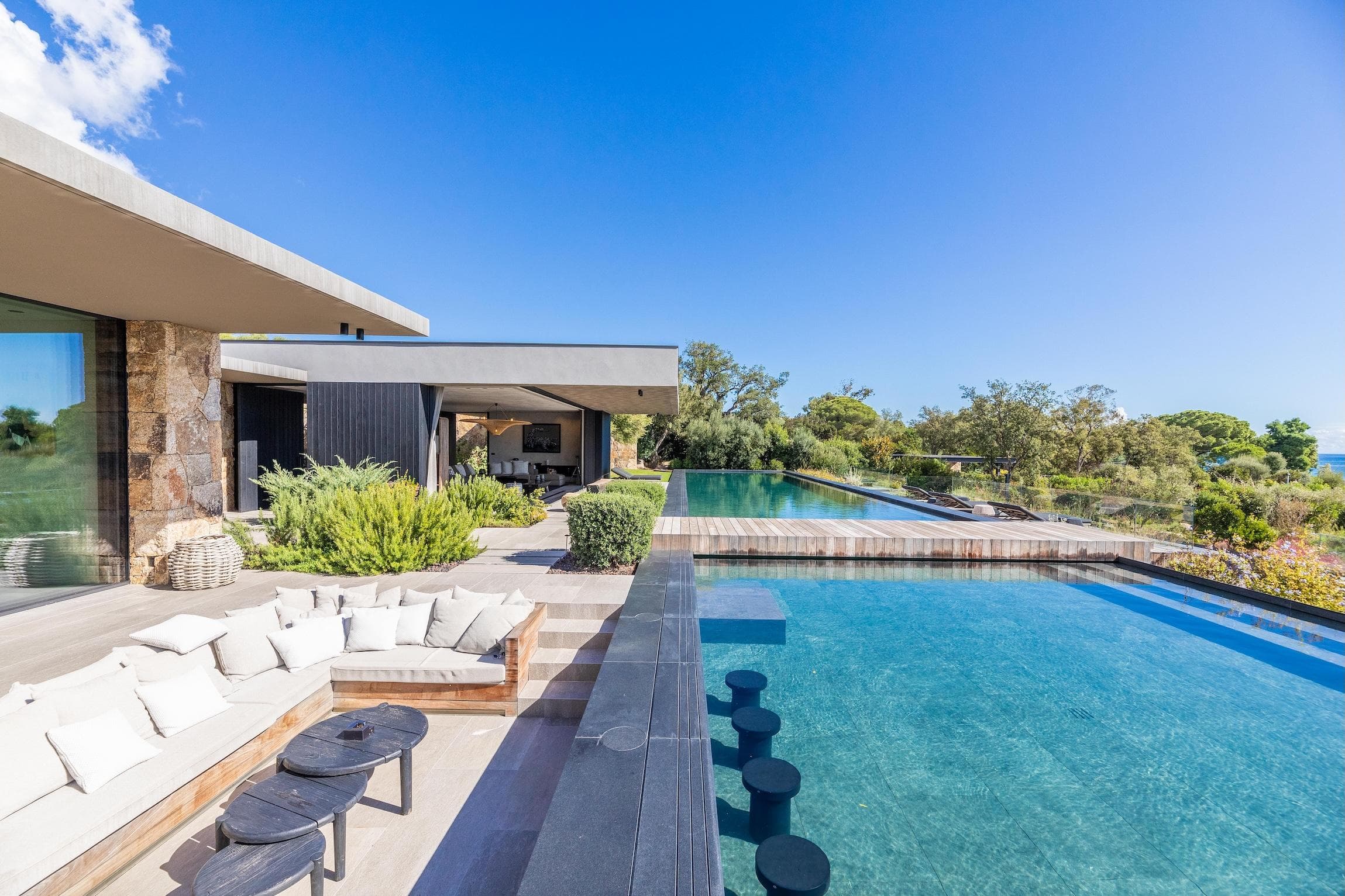 Modern house with large glass windows, a sunken outdoor seating area with white cushions, and an infinity pool, surrounded by greenery and under a clear blue sky.